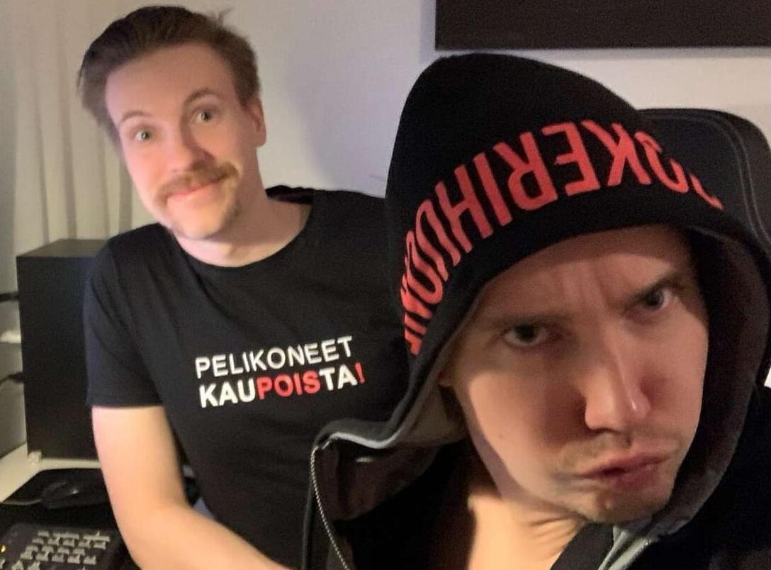 Mies huppu päässä ja duckface naamalla, takana toinen hymyilee paidassa jossa lukee "pelikoneet pois kaupoista"