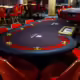 Paljon pokeripöytiä salissa. Pöydän pinnat ovat tummia ja pöytien tuolit ovat punaisia.Kokolattiamatto on myös tumma.