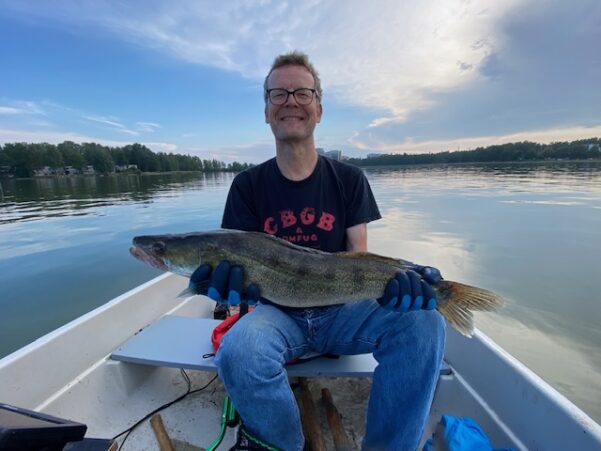 Jorma Vuoksenmaa istuu veneessä hymy huulilla iso kuha kädessän. Taustalla lähes pilveetön taivas ja tyyni meri