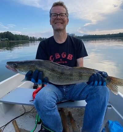 Jorma Vuoksenmaa istuu veneessä hymy huulilla iso kuha kädessän. Taustalla lähes pilveetön taivas ja tyyni meri