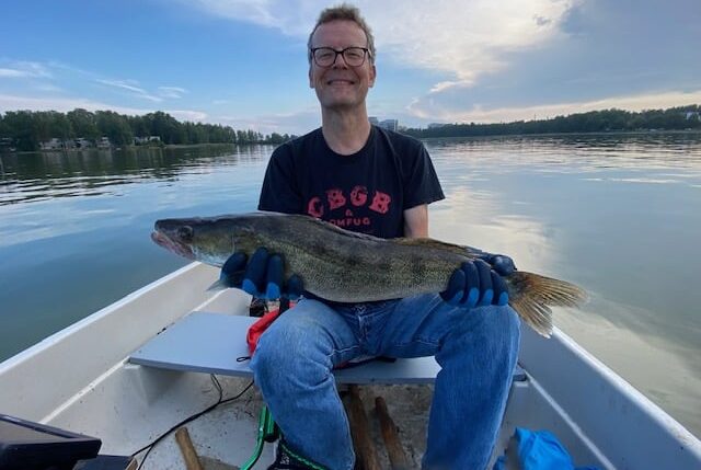 Jorma Vuoksenmaa istuu veneessä hymy huulilla iso kuha kädessän. Taustalla lähes pilveetön taivas ja tyyni meri