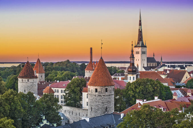 Kuvassa Tallinnan vanhan kaupungin punakattoisia torneja ja vihreitä puiden latvoja. Taustalla näkyy auringonlasku ja meri.
