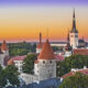 Tallinnan siluetti ja vanhan kaupungin punakattoisia torneja ja vihreitä puiden latvoja. Taustalla näkyy auringonlasku ja meri.