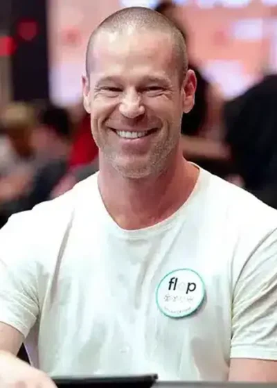 Patrik Antonius istuu pokeripöydässä ja naureskelee. Hänellä on päällään valkoinen t-paita ja hän räplää pelimerkkejä pöydällä.