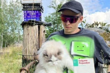 Mies istuu aurinkolasit päässä lippis ja aurinkolasit. Miehellä on sylissään kissa. Takana puun päällä on kasa erilaisia kirjoja.