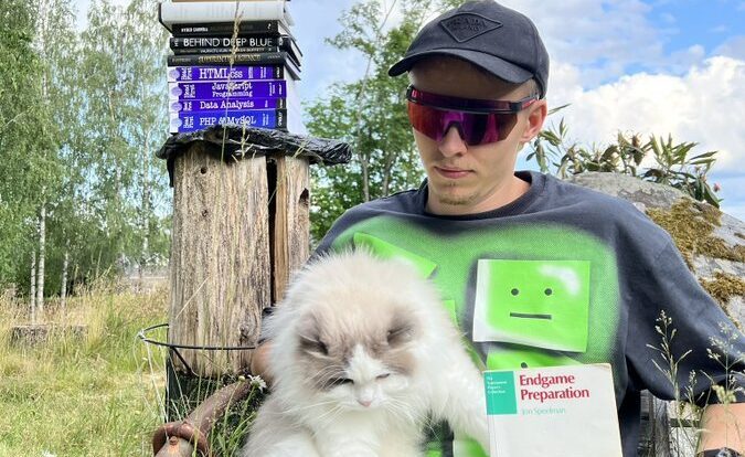 Mies istuu aurinkolasit päässä lippis ja aurinkolasit. Miehellä on sylissään kissa. Takana puun päällä on kasa erilaisia kirjoja.