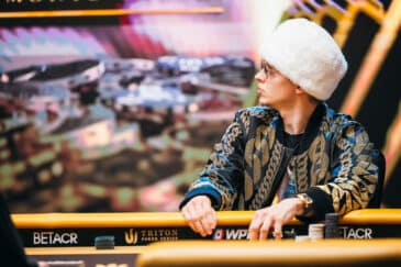 Mies valkeassa karvahatussa katsoo sivulle. Mies istuu pokeripöydän ääressä.