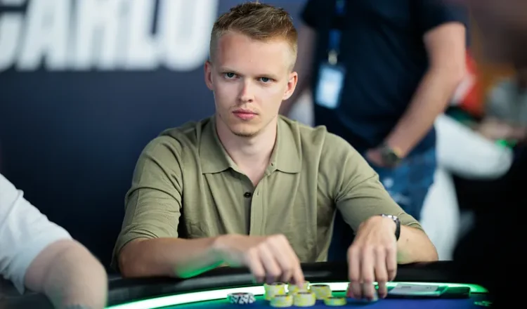 Mies istuu pokeripöydässä. Hänellä on päällään vaaleanruskea kauluspaita. Pöydällä miehen edessä on erivärisiä pelimerkkejä.