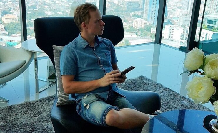 Mikko Rahtu istuu nojatuolissa. Hänellä oikea jalka vasemman jalan päällä ja kädessään puhelin. Taustalla avautuu upeat maisemat