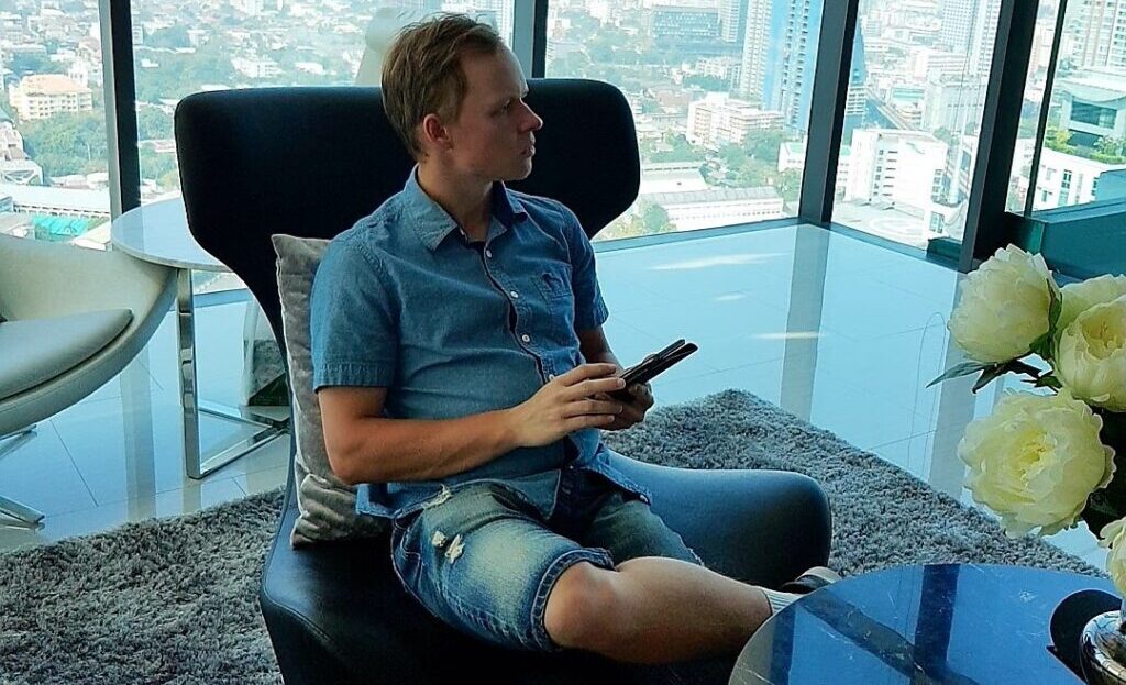 Mikko Rahtu istuu nojatuolissa. Hänellä oikea jalka vasemman jalan päällä ja kädessään puhelin. Taustalla avautuu upeat maisemat