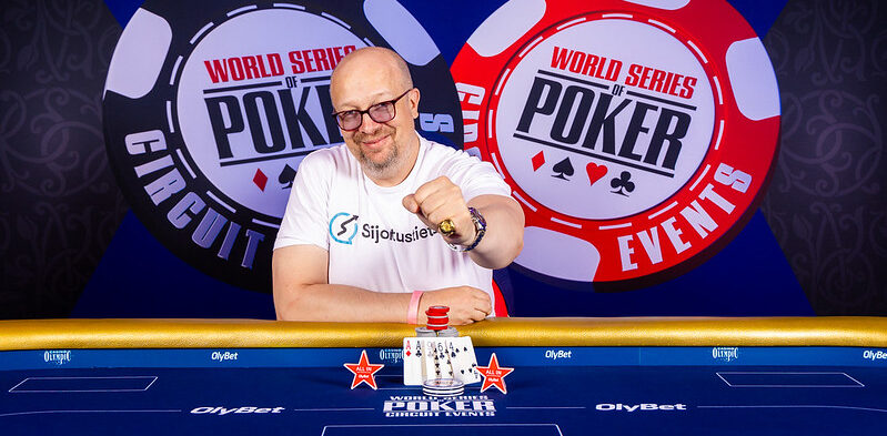 Aki Pyysing hymyilee voittajakuvassa Tallinnan WSOPC:ssa. Pyysingillä on sormessaan voittosormus ja päällään valkoinen t-paita.