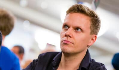 Mies katsoo vasemmalle. Hänellä on lyhyet hiukset ja musta kauluspaita. Vieressä näkyy sinipaitainen mies. Tausta on vaaleaa.