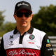 Kimi Räikkönen seisoo päässään musta lippis. Hänellä on päällään Alfa-Romeon pikeepaita. Taustalla näkyy puita ja metalliaita.