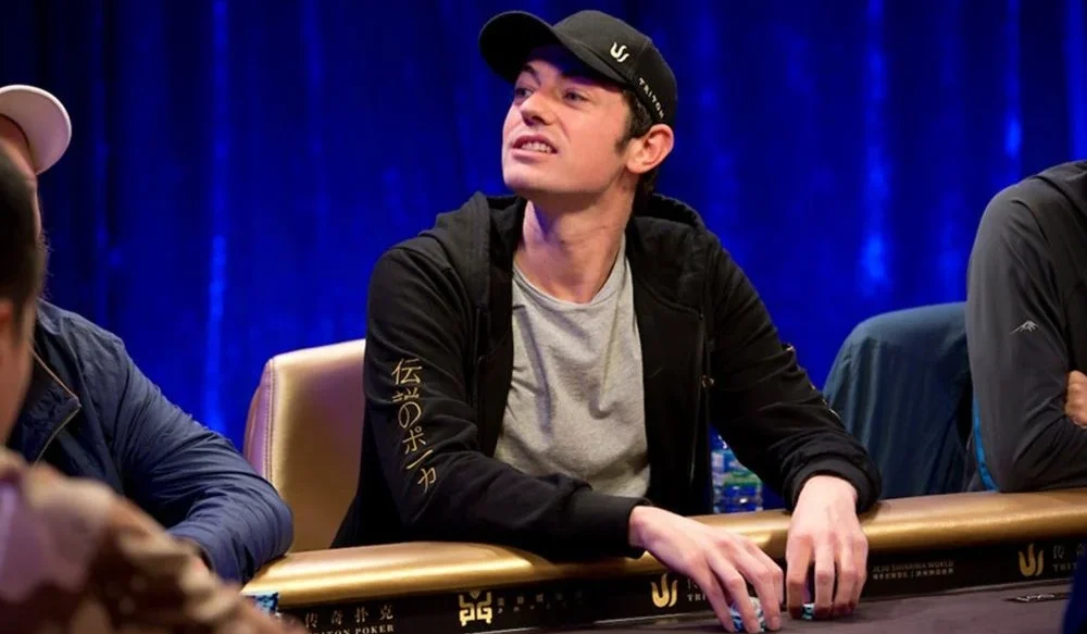 Tom Dwan istuu pokeripöydässä Triton Poker Serieksen turnauksessa. Hänellä on päällään huppari, joka on auki ja musta lippis.