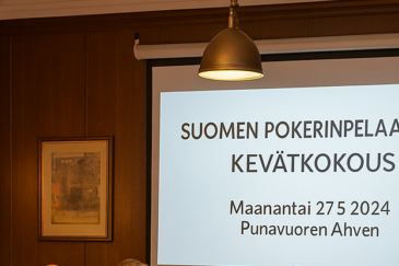 Valkokankaalla lukee Suomen pokerinpelaajat ry kevätkokous. Vasemmalla puolella on ruskeaa seinää, jossa on muutamia tauluja.