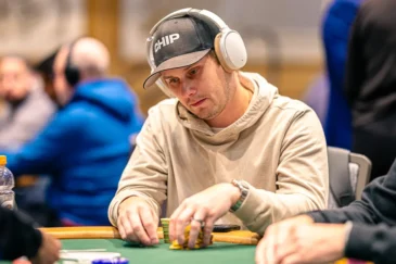 Mies on pokeripöydässä. Päässään miehellä on musta lippis, jossa lukee valkoisella tekstiä. Hän pitelee käsissään pelimerkkejä.