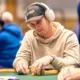 Mies on pokeripöydässä. Päässään miehellä on musta lippis, jossa lukee valkoisella tekstiä. Hän pitelee käsissään pelimerkkejä.