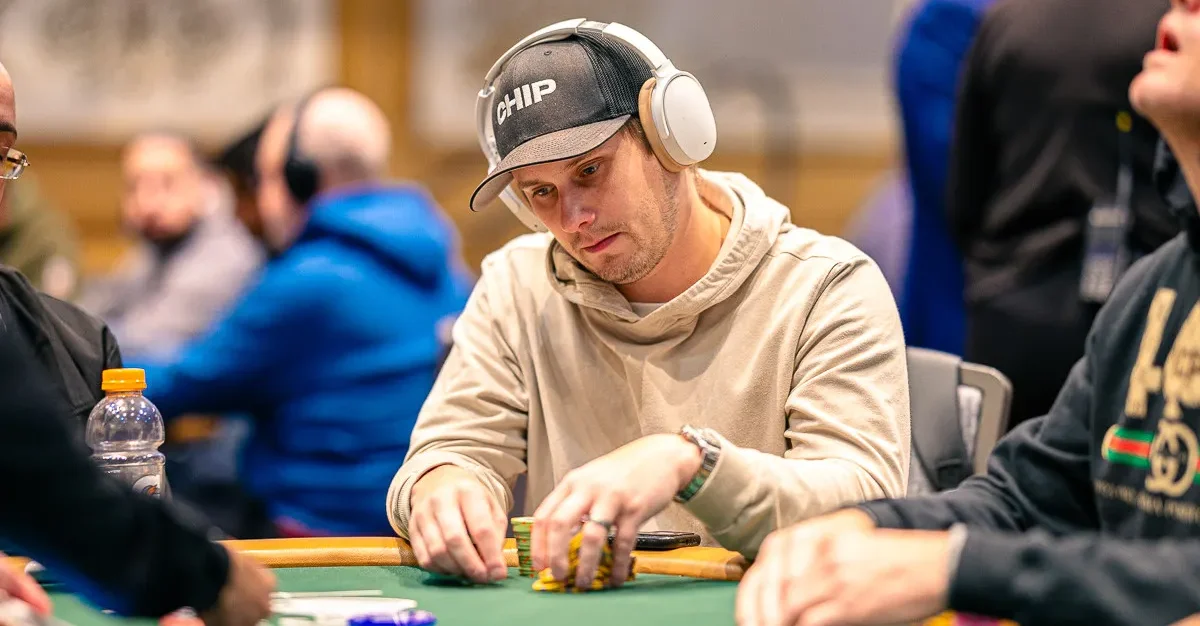 Mies on pokeripöydässä. Päässään miehellä on musta lippis, jossa lukee valkoisella tekstiä. Hän pitelee käsissään pelimerkkejä.