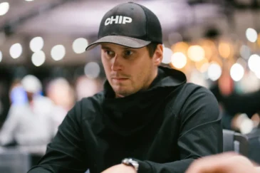 Mies istuu pokeripöydässä. Hänellä on päällään musta paita ja musta lippis. Miehellä on vasemmassa kädessään hopeinen kello.