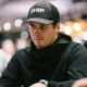 Mies istuu pokeripöydässä. Hänellä on päällään musta paita ja musta lippis. Miehellä on vasemmassa kädessään hopeinen kello.