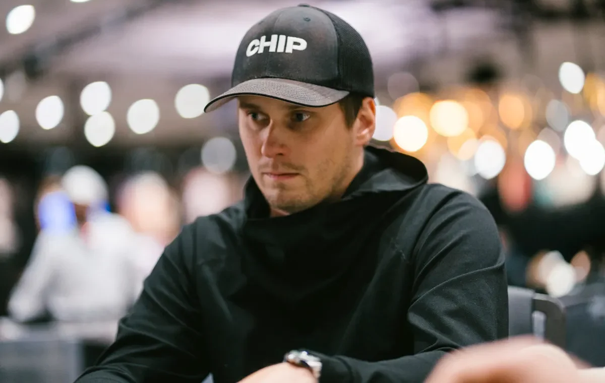 Mies istuu pokeripöydässä. Hänellä on päällään musta paita ja musta lippis. Miehellä on vasemmassa kädessään hopeinen kello.