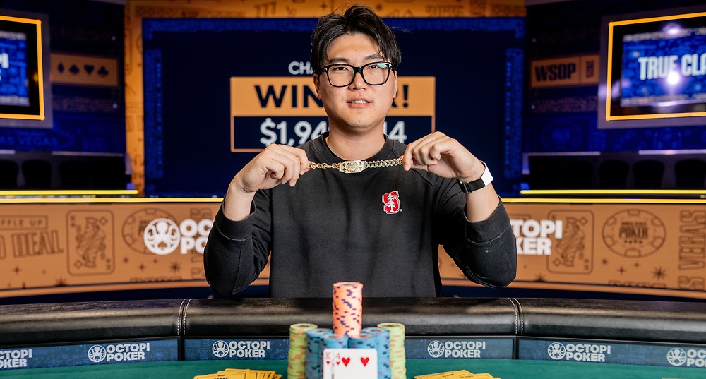 Mies istuu pokeripöydässä. Hänellä on päällään musta paita. Käsissään hän pitää kultaista ranneketta. Tausta on vaalean ruskeaa.
