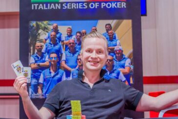 Mies istuu pokeripöydässä ja hymyilee leveästi. Hänellä on toisessa kädessään pelikortteja. Takana näkyy puuseinä ja juliste.