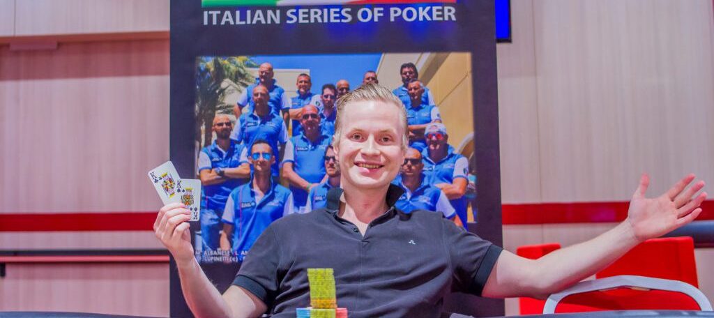 Mies istuu pokeripöydässä ja hymyilee leveästi. Hänellä on toisessa kädessään pelikortteja. Takana näkyy puuseinä ja juliste.