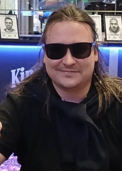 Pitkähiuksinen mies istuu pokeripöydässä. Hänellä on päällään musta t-paita sekä mustat aurinkolasit. Hän näyttää myös peukkua.