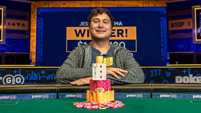 Mies istuu pokeripöydässä päällään harmaa huppari. Hän hymyilee ja hänen edessään on keltaisia ja pinkkejä pelimerkkejä.