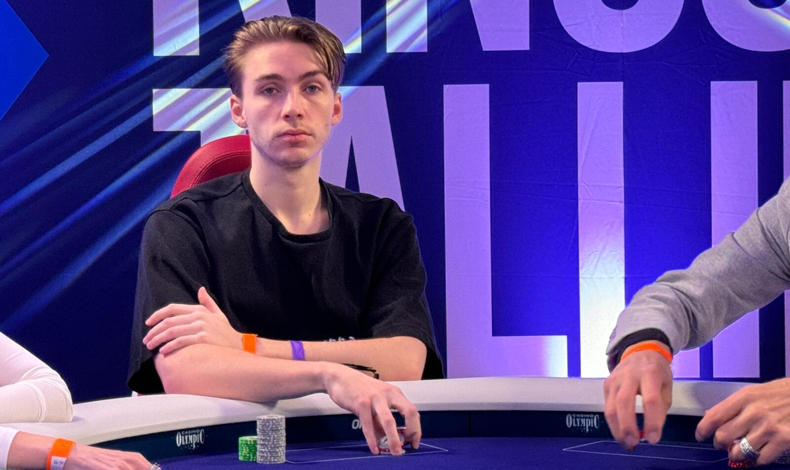 Mies istuu pokeripöydässä. Hänellä on päällään musta t-paita. Taustalla on sininen mainos, jossa on on valkoisella tekstiä.