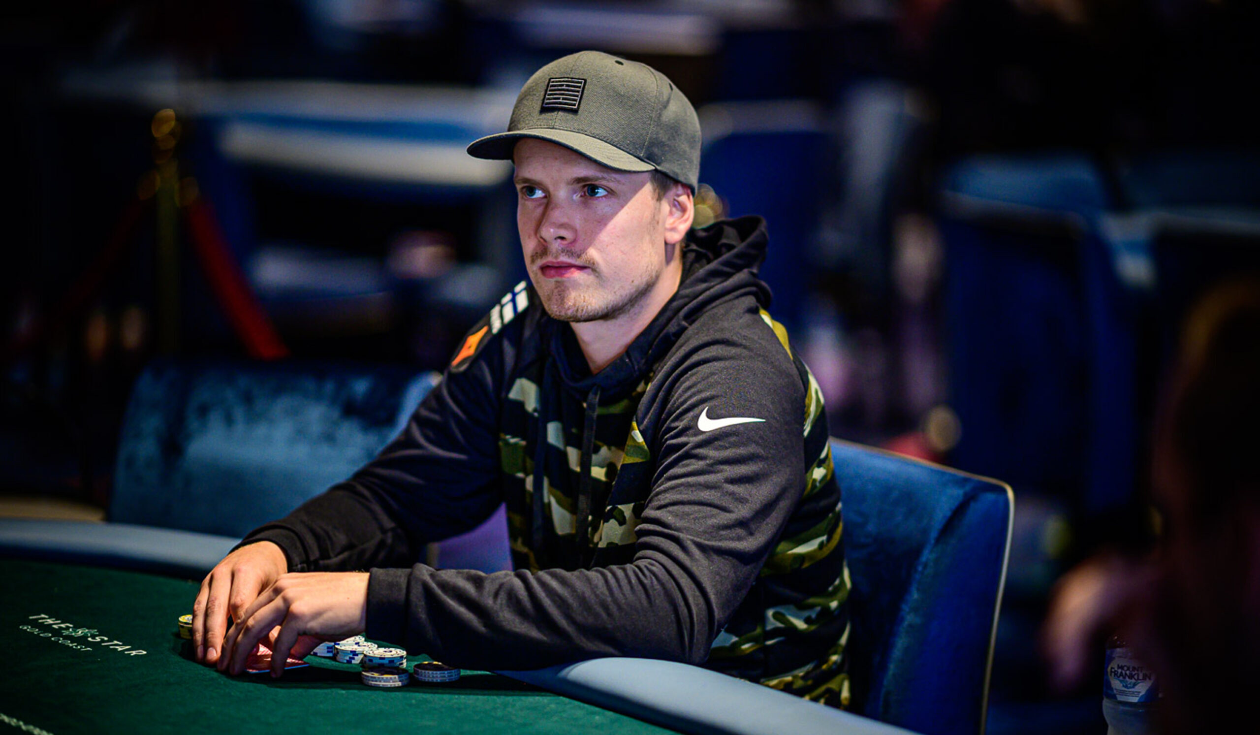 Mies istuu pokeripöydässä sinisellä penkillä. Hänellä on päällään musta Niken huppari. Päässään hänellä on tumma lippis.