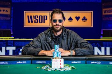 Mies istuu pokeripöydässä ja hymyilee. Hänellä on päällään musta takki ja päässään aurinkolasit. Kauempana taustalla lukee WSOP.