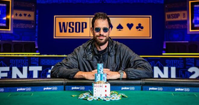 Mies istuu pokeripöydässä ja hymyilee. Hänellä on päällään musta takki ja päässään aurinkolasit. Kauempana taustalla lukee WSOP.