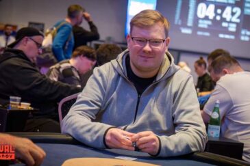 Mies istuu pokeripöydässä. Hänellä on päällään harmaa huppari.Käsissään mies pitää pelimerkkejä. Taustalla istuu muita ihmisiä.