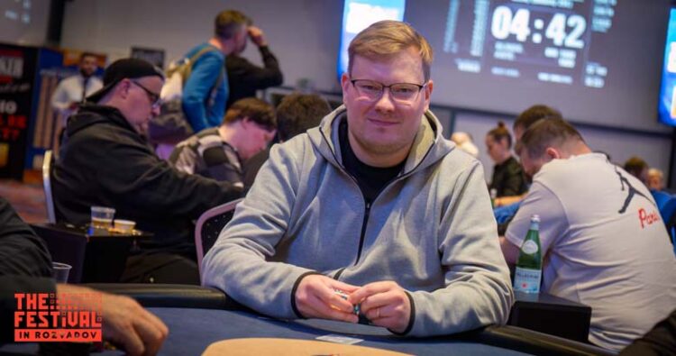 Mies istuu pokeripöydässä. Hänellä on päällään harmaa huppari.Käsissään mies pitää pelimerkkejä. Taustalla istuu muita ihmisiä.