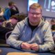 Mies istuu pokeripöydässä. Hänellä on päällään harmaa huppari.Käsissään mies pitää pelimerkkejä. Taustalla istuu muita ihmisiä.