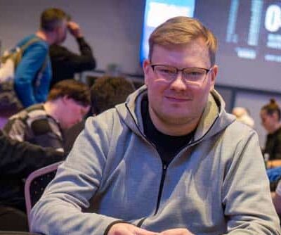 Mies istuu pokeripöydässä. Hänellä on päällään harmaa huppari.Käsissään mies pitää pelimerkkejä. Taustalla istuu muita ihmisiä.
