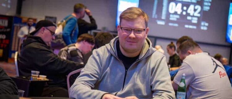 Mies istuu pokeripöydässä. Hänellä on päällään harmaa huppari.Käsissään mies pitää pelimerkkejä. Taustalla istuu muita ihmisiä.