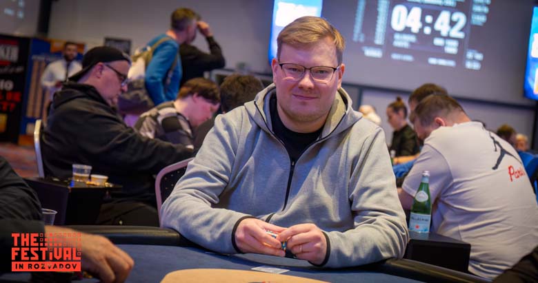 Mies istuu pokeripöydässä. Hänellä on päällään harmaa huppari.Käsissään mies pitää pelimerkkejä. Taustalla istuu muita ihmisiä.