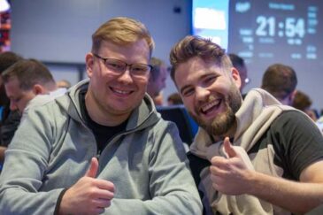 Kuvassa on kaksi iloista miestä, toisella on harmaa paita, ja toisella vaalea paita. Taustalla näkyy sinisellä näytöllä ajastin.