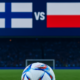 Jalkapallo vihreällä nurmella. Pallon takana kauempana on jalkapallomaali ja katsomo. Myös Suomen ja Puolan liput ovat kuvassa.