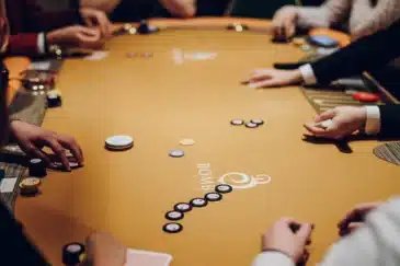 Kuvassa on keltapintainen pokeripöytä. Pöydällä näkyy paljon käsiä. Pöydällä on myös erilaisia pelimerkkejä ja pelikortteja.