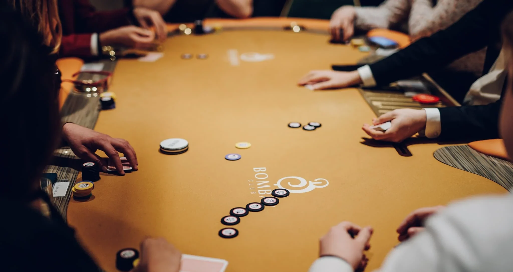 Kuvassa on keltapintainen pokeripöytä. Pöydällä näkyy paljon käsiä. Pöydällä on myös erilaisia pelimerkkejä ja pelikortteja.
