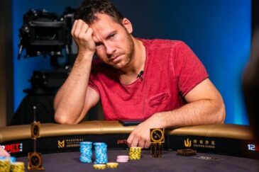 Mies istuu pokeripöydässä päällään punainen paita. Hän nojaa kädellään päähänsä ja hänen ilme on mietteliäs. Pöydällä on pelimerkkejä.