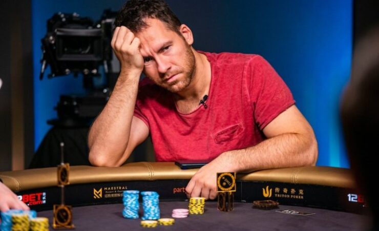 Mies istuu pokeripöydässä päällään punainen paita. Hän nojaa kädellään päähänsä ja hänen ilme on mietteliäs. Pöydällä on pelimerkkejä.