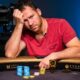 Mies istuu pokeripöydässä päällään punainen paita. Hän nojaa kädellään päähänsä ja hänen ilme on mietteliäs. Pöydällä on pelimerkkejä.