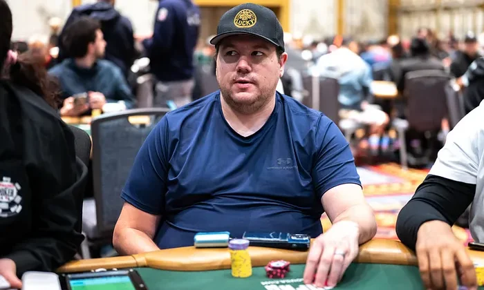 Mies istuu pokeripöydässä. Hänellä on päällään sininen paita ja musta lippis. Pöydällä on erivärisiä pelimerkkejä.