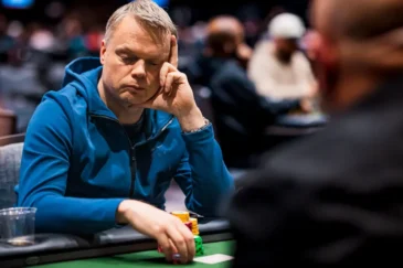 Mies istuu pokeripöydässä. Hänellä on mietteliäs ilme ja hän nojaa käteensä. Miehellä on toisessa kädessään pelimerkkejä.