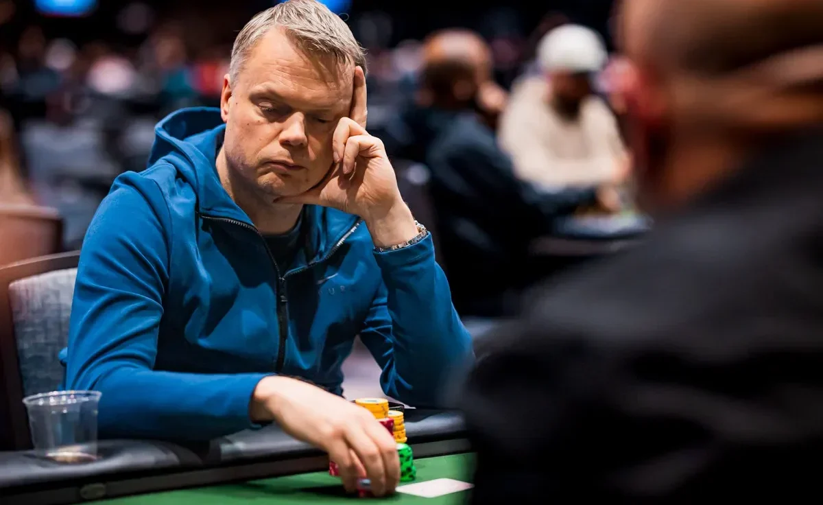 Mies istuu pokeripöydässä. Hänellä on mietteliäs ilme ja hän nojaa käteensä. Miehellä on toisessa kädessään pelimerkkejä.